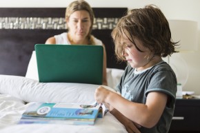 5 year old boy looking at book in hotel room as mom works on laptop