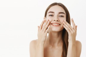 Waist-up shot of charming relaxed and gentle young woman making cosmetological procedure applying facial cream on face with fingers and smiling broadly feeling perfect, taking care of skin