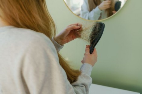 A young woman gently pulls loose hair from a hairbrush. The black brush is filled with fallen hair, emphasizing hair loss and daily hair care routine.
